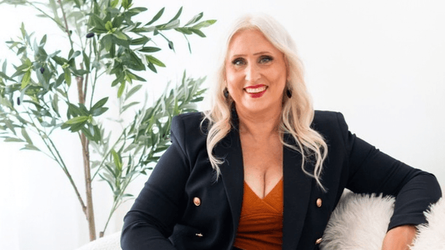Smiling woman in a black blazer poses on a sofa near a plant