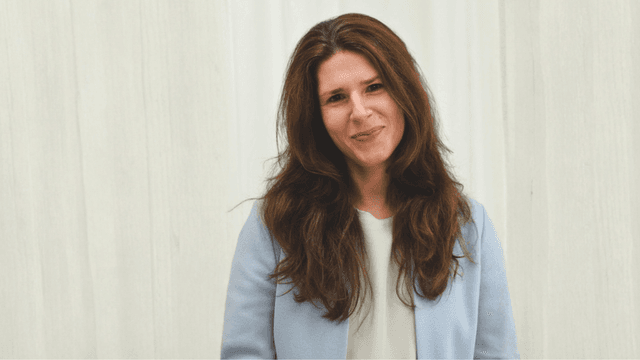 Woman with long brown hair in a light blue blazer stands smiling against a white background