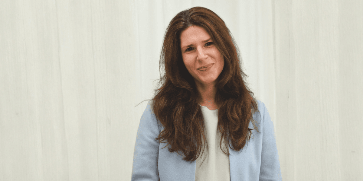 Woman with long brown hair in a light blue blazer stands smiling against a white background