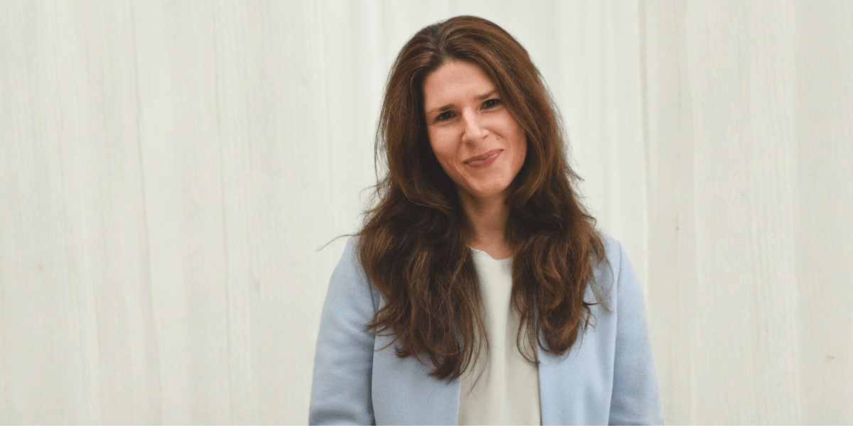 Woman with long brown hair in a light blue blazer stands smiling against a white background