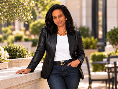 A woman in a blazer and jeans poses confidently outdoors with greenery around her