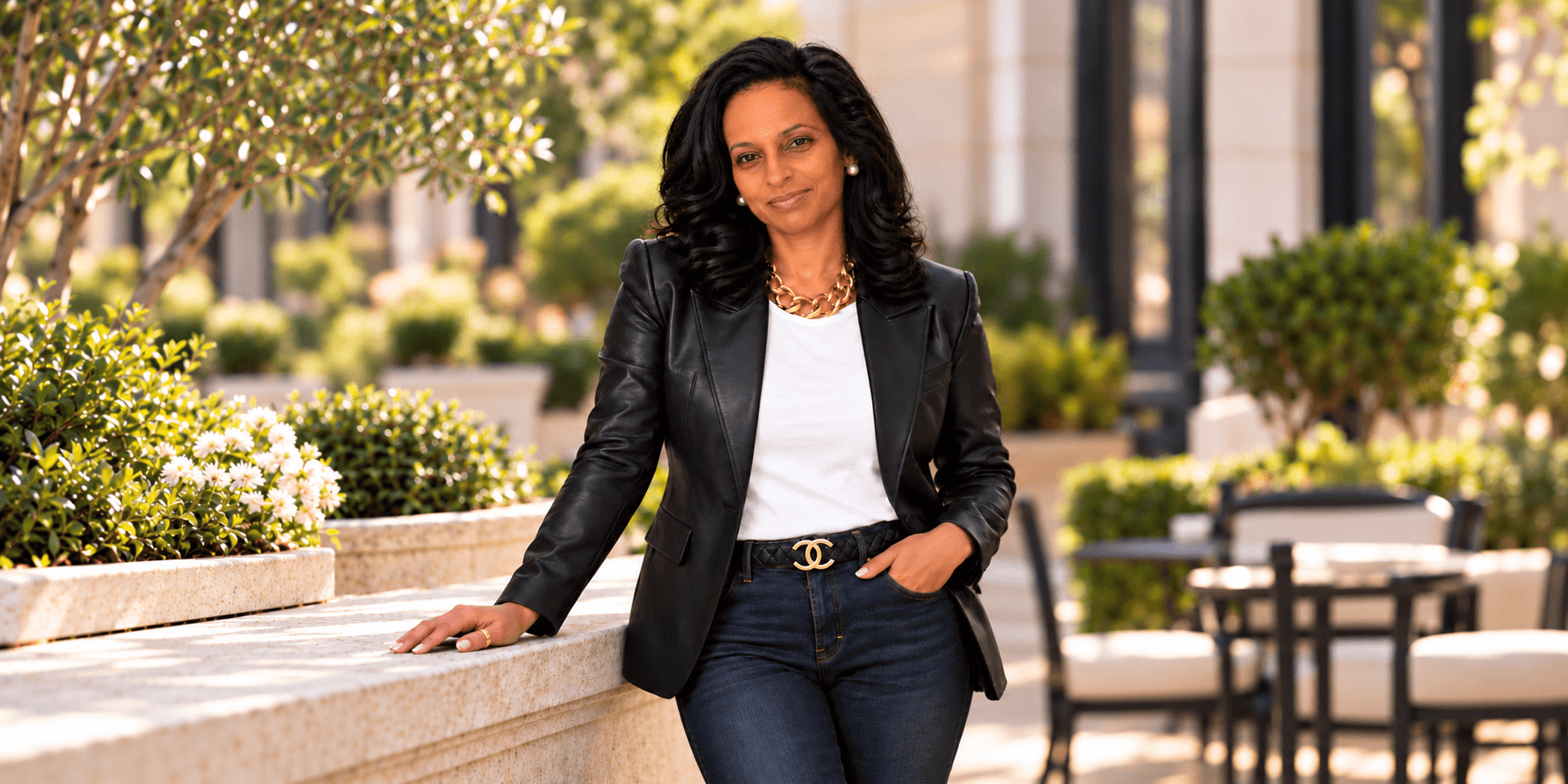 A woman in a blazer and jeans poses confidently outdoors with greenery around her