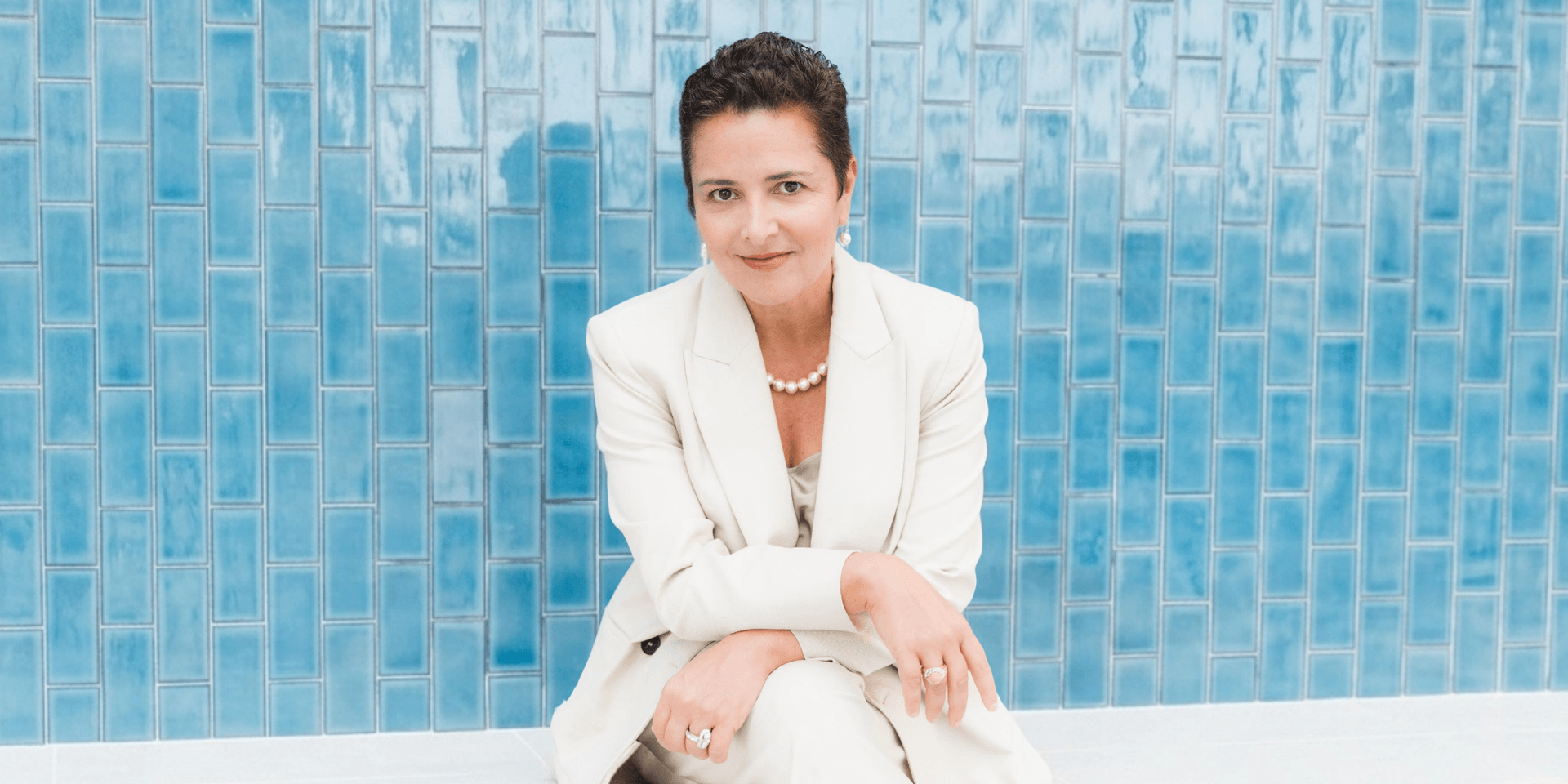 A woman in a cream suit sits against a blue tiled wall, smiling gently
