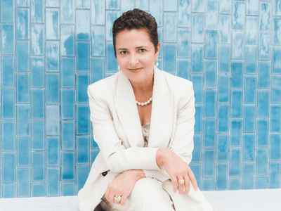 A woman in a cream suit sits against a blue tiled wall, smiling gently
