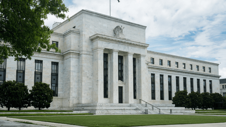 Federal Reserve building with marble facade, columns, and flag against a cloudy sky