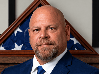 Man with a shaved head in a suit, standing in front of a flag display