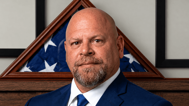 Man with a shaved head in a suit, standing in front of a flag display