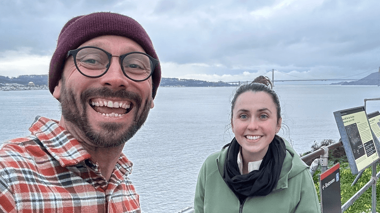 A man and woman smiling at a scenic viewpoint, with the Golden Gate Bridge in the background, enjoying the view.