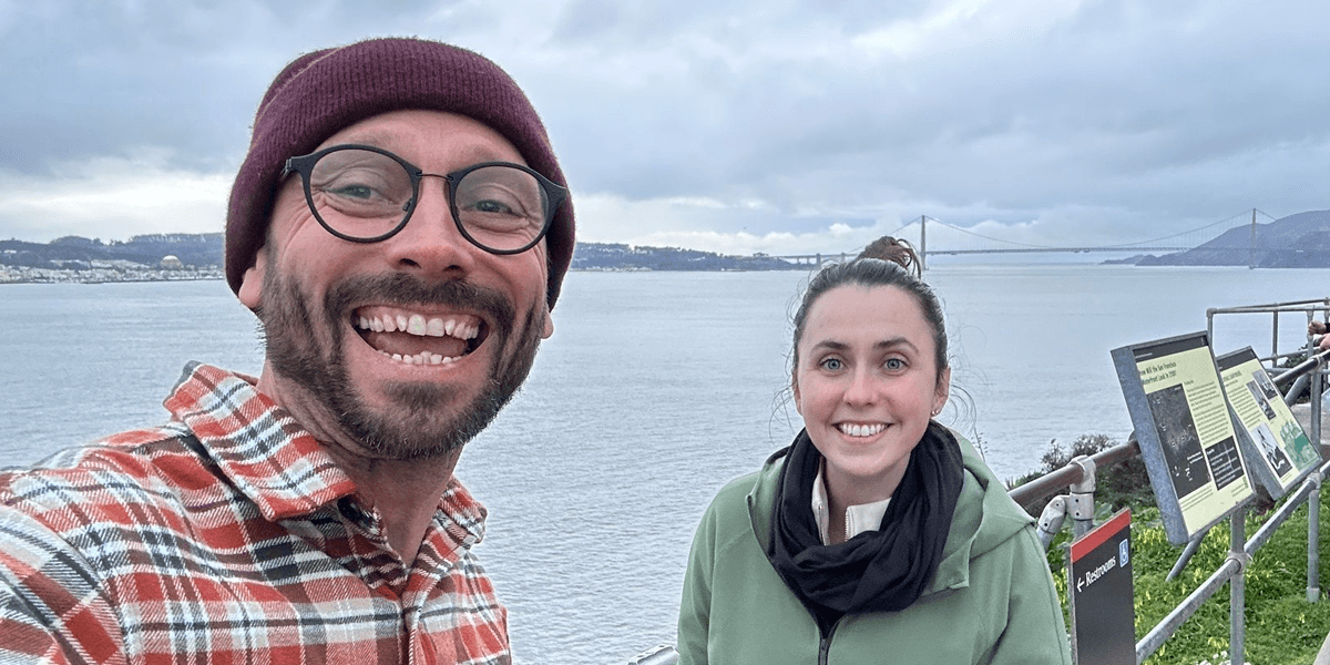 A man and woman smiling at a scenic viewpoint, with the Golden Gate Bridge in the background, enjoying the view.