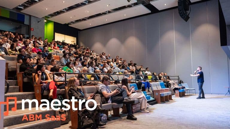 Audience watching a presenter in a modern auditorium, with "maestro ABM SaaS" logo in the foreground. The tone is professional and engaging.