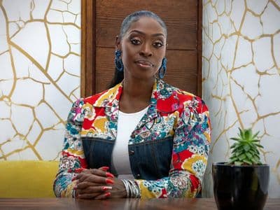 Dilys Sillah seated at a table wearing a colorful jacket in a contemporary office setting.