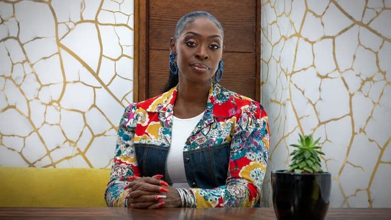 Dilys Sillah seated at a table wearing a colorful jacket in a contemporary office setting.