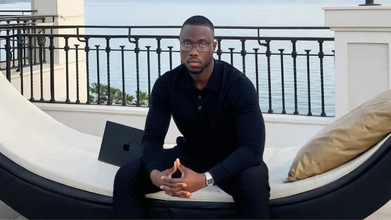 A man in a black polo shirt sits outdoors by the ocean with an Apple laptop, reflecting on sales systems.