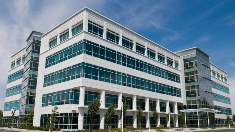 Modern glass office building with contemporary architecture, reflective windows, and landscaped entrance under a clear blue sky.