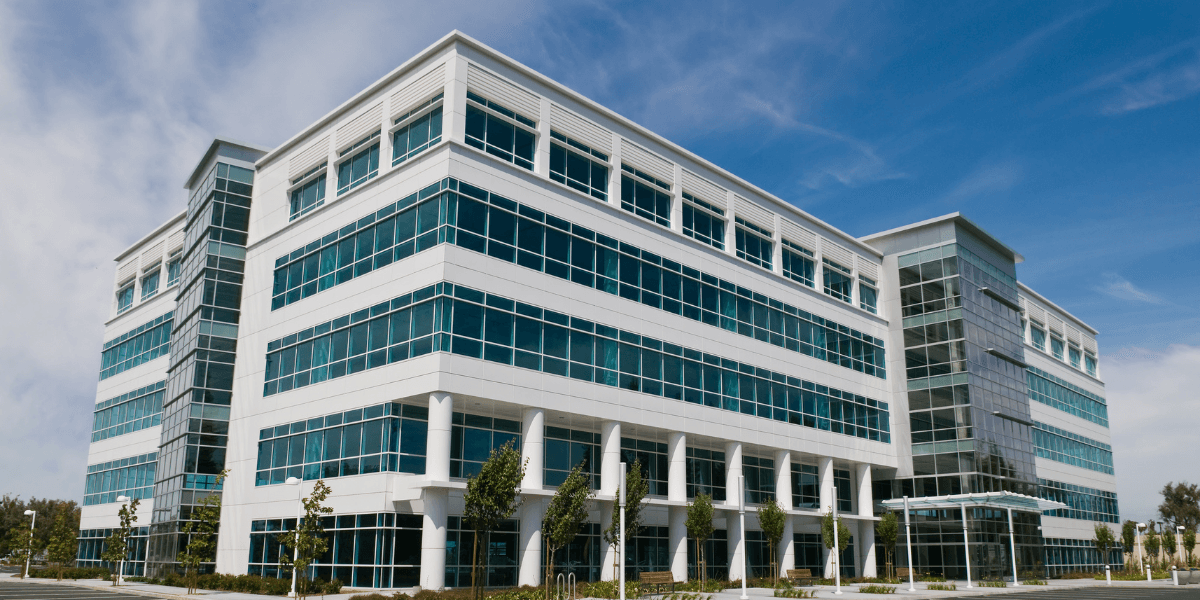 Modern glass office building with contemporary architecture, reflective windows, and landscaped entrance under a clear blue sky.