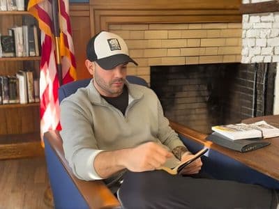 Jason Xavier Duffy seated in a library reading from a tablet beside an American flag.