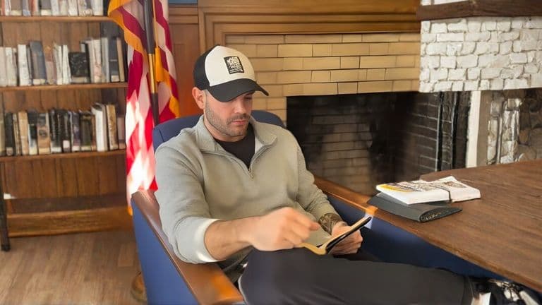Jason Xavier Duffy seated in a library reading from a tablet beside an American flag.