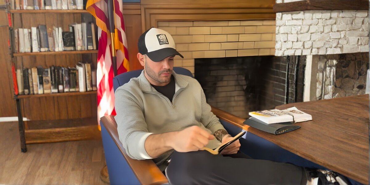 Jason Xavier Duffy seated in a library reading from a tablet beside an American flag.