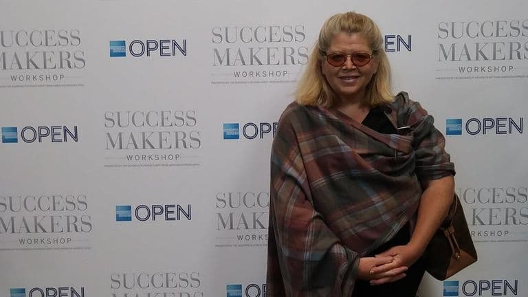Janet Walker posing at Success Makers Workshop event backdrop wearing plaid shawl and sunglasses.