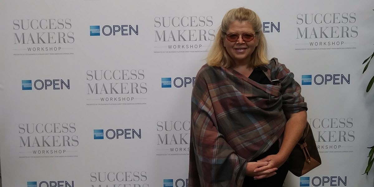 Janet Walker posing at Success Makers Workshop event backdrop wearing plaid shawl and sunglasses.