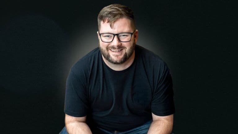 Will Craig smiling in glasses and a black shirt against a dark background.