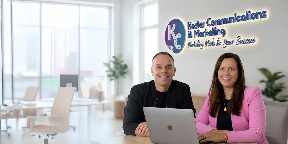 Two people seated at a desk in a modern office, smiling at the camera. A company logo and slogan are visible on the wall behind them.