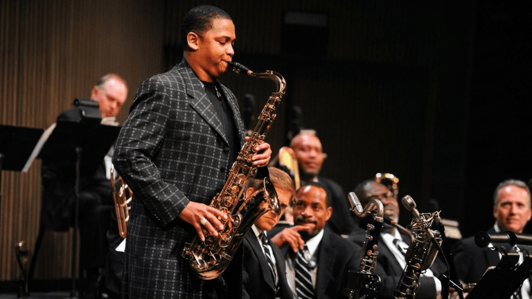 Jazz saxophonist performing solo on stage with orchestra seated behind him.