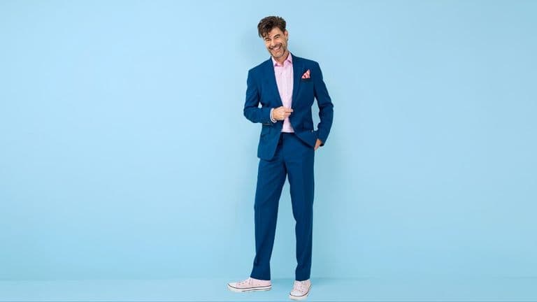 A man in a blue suit with a pink shirt and pocket square stands against a light blue background, smiling. He exudes a casual and confident vibe.