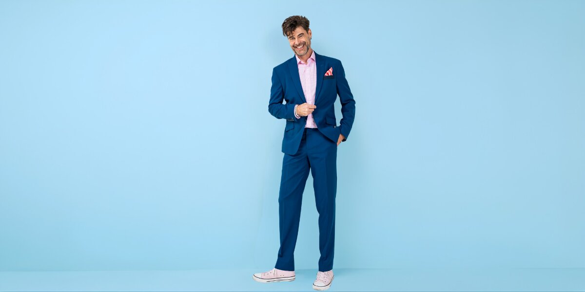 A man in a blue suit with a pink shirt and pocket square stands against a light blue background, smiling. He exudes a casual and confident vibe.