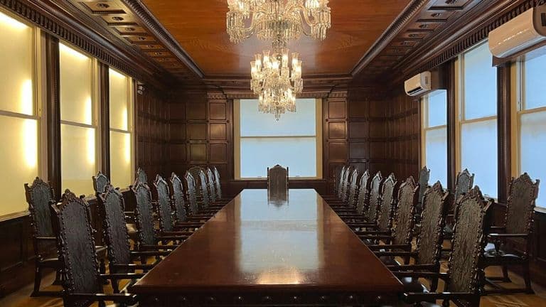 Large, elegant boardroom with a long, polished wooden table. Ornate chairs line both sides. Two chandeliers hang overhead, casting a warm glow.