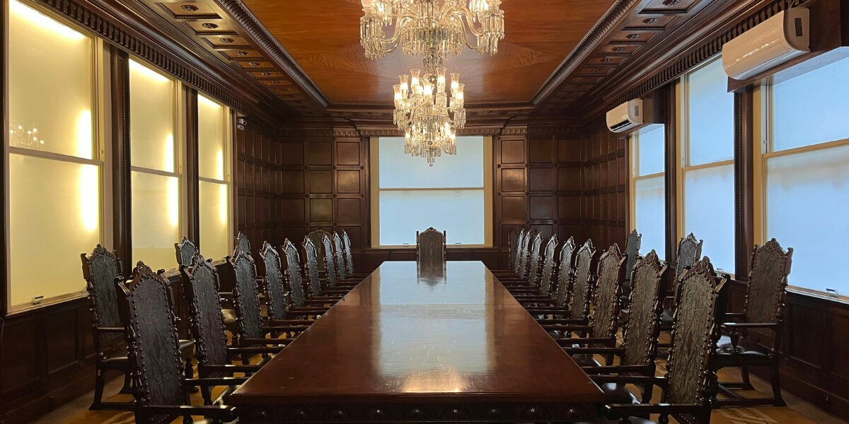 Large, elegant boardroom with a long, polished wooden table. Ornate chairs line both sides. Two chandeliers hang overhead, casting a warm glow.