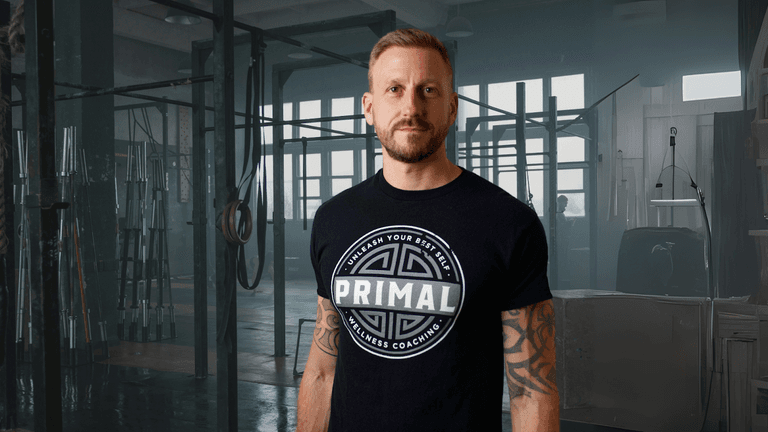 A man wearing a "PRIMAL Wellness Coaching" t-shirt stands in a gym, surrounded by fitness equipment, looking confidently at the camera.