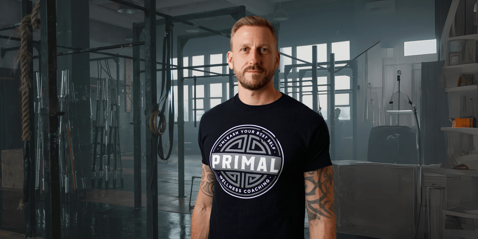 A man wearing a "PRIMAL Wellness Coaching" t-shirt stands in a gym, surrounded by fitness equipment, looking confidently at the camera.
