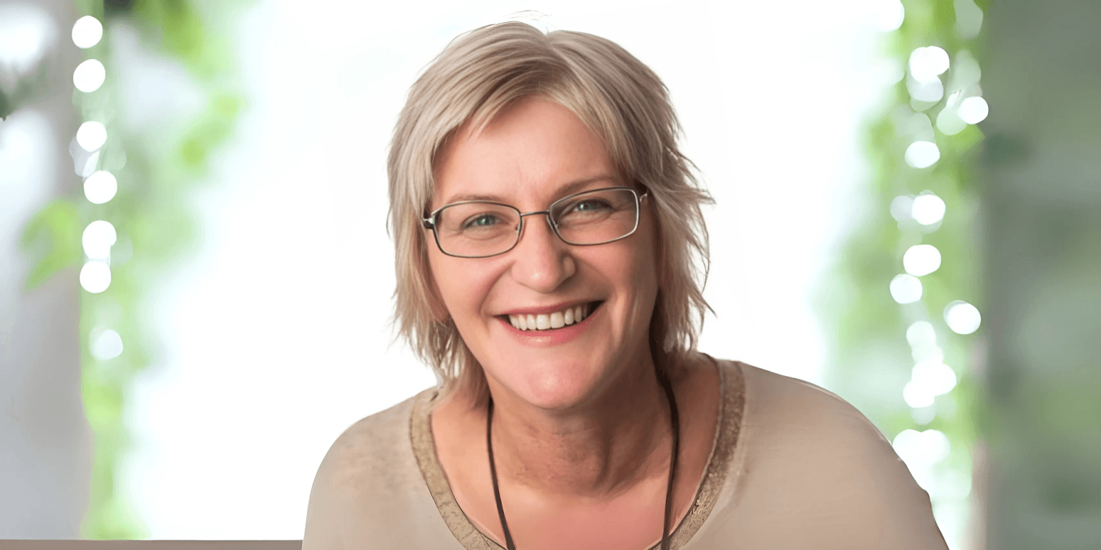 Smiling woman with short blonde hair and glasses, wearing a light-colored shirt, with a soft, blurred background of greenery and bokeh lights, radiating a warm and welcoming demeanor.