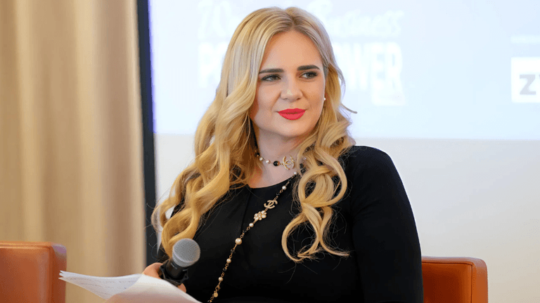 Blonde woman in a black dress with a Chanel necklace, holding a microphone and looking off to the side during a panel discussion.