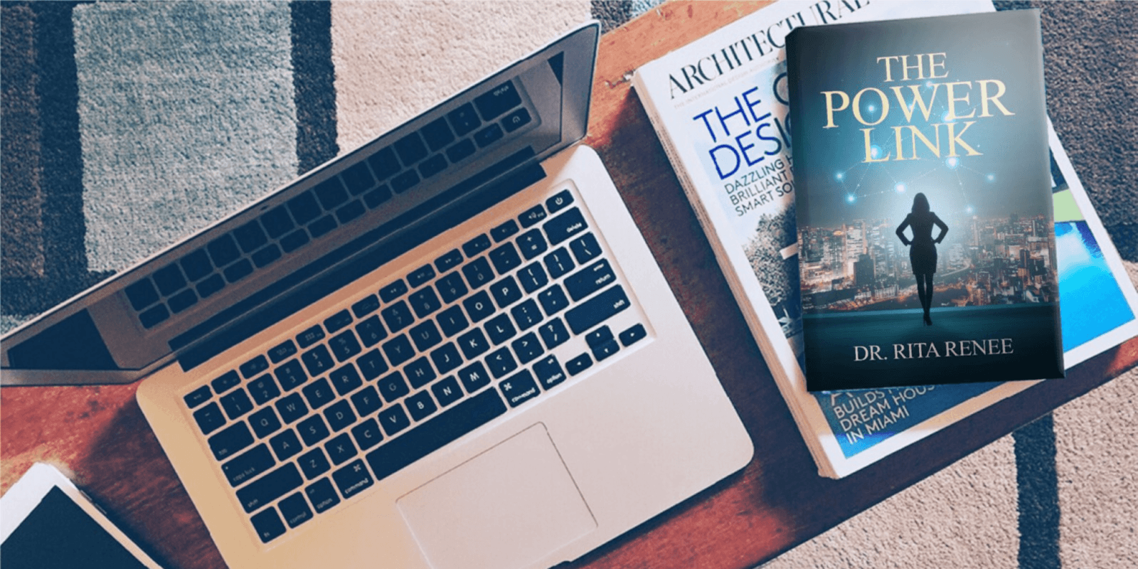 Laptop with architectural magazine and book titled 'The Power Link' by Dr. Rita Renee on a patterned rug, showcasing a modern business setup with a focus on professional development.